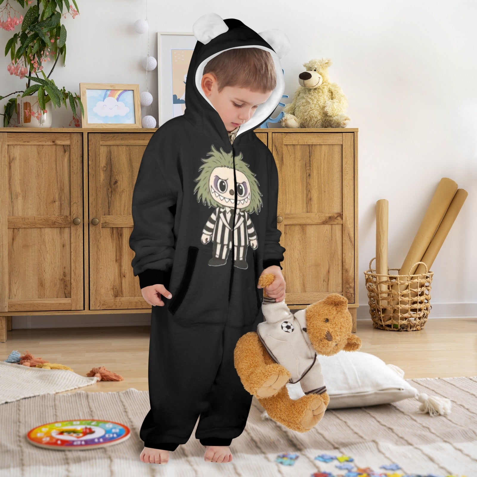Child wearing a black onesie with a cartoon design, holding a teddy bear in a room with wooden furniture and toys.