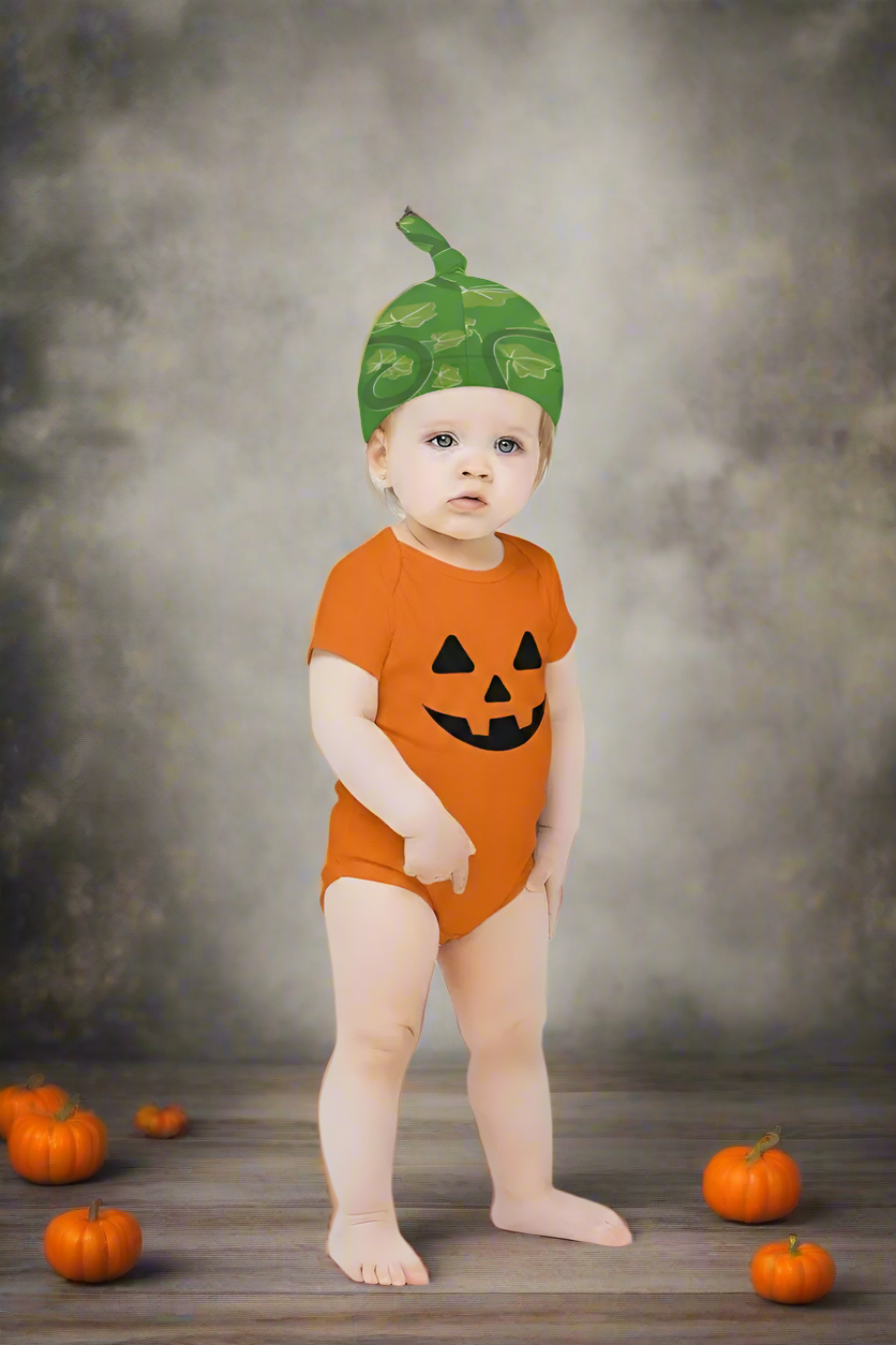 Baby wearing an orange onesie with a pumpkin face and green hat on a light gray background